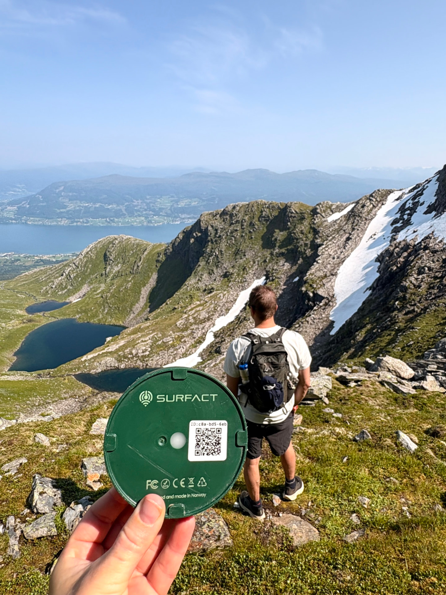 Emma Smart Tracker held up on a ridge above a fjord with mountain lakes below