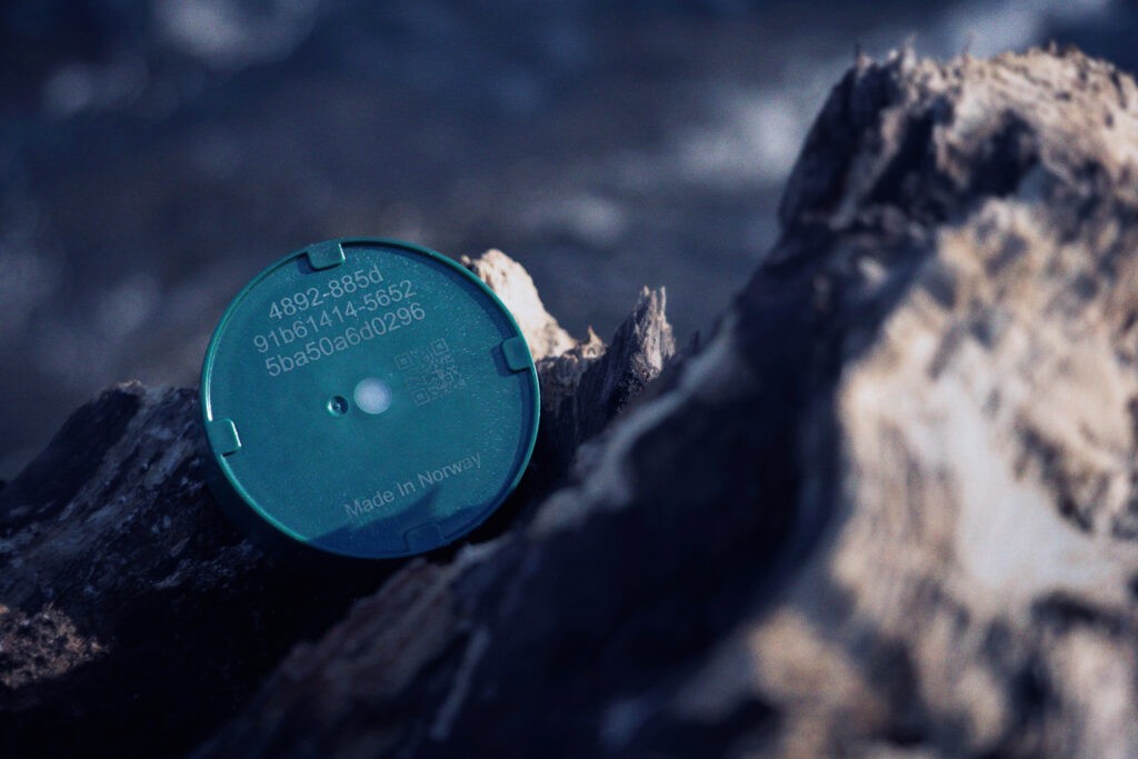 Circular green Emma IoT tracker resting on a dark rock, marked Made in Norway