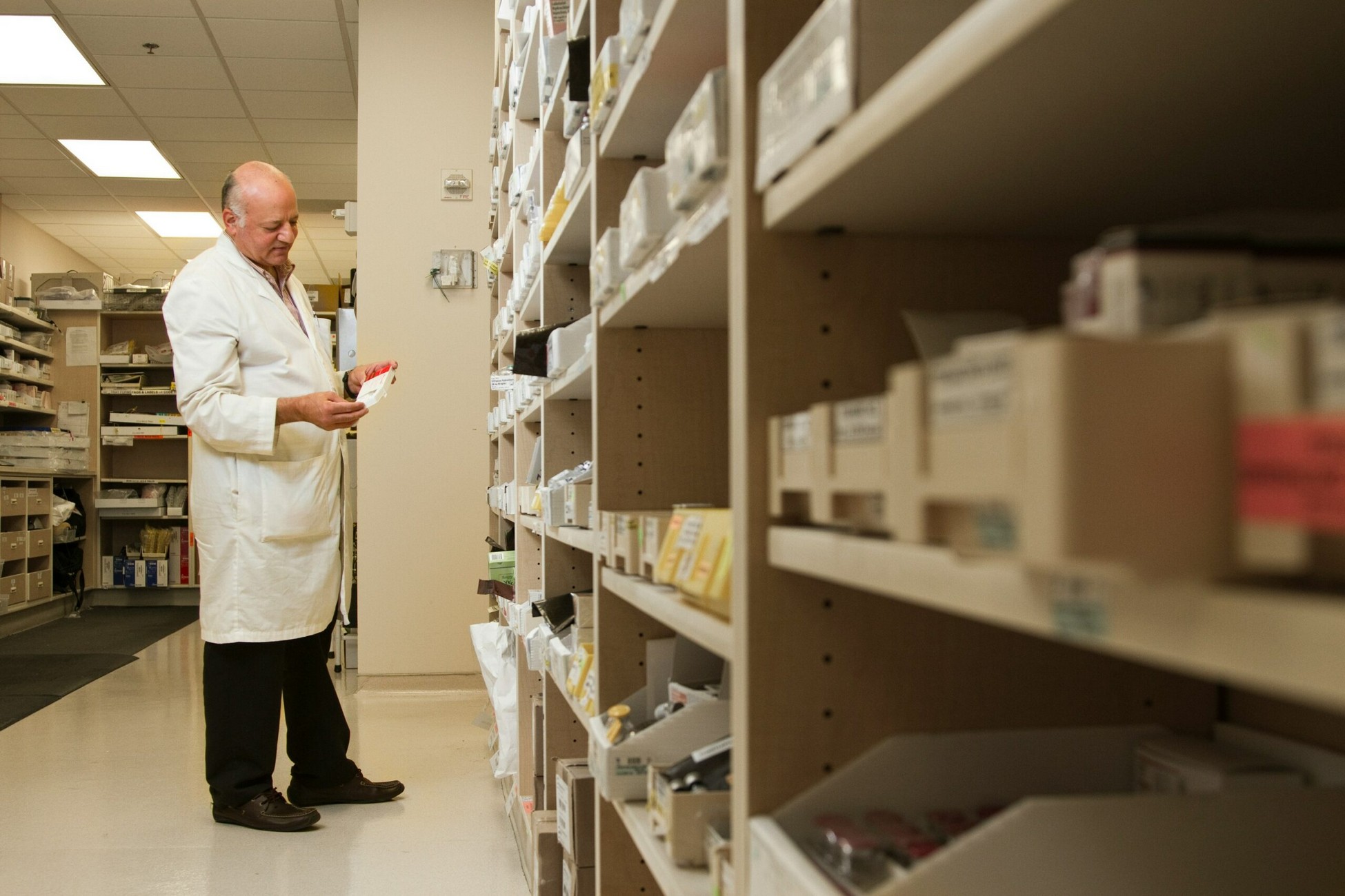 Pharmacist in a white lab coat inspecting a medication box in a pharmacy aisle