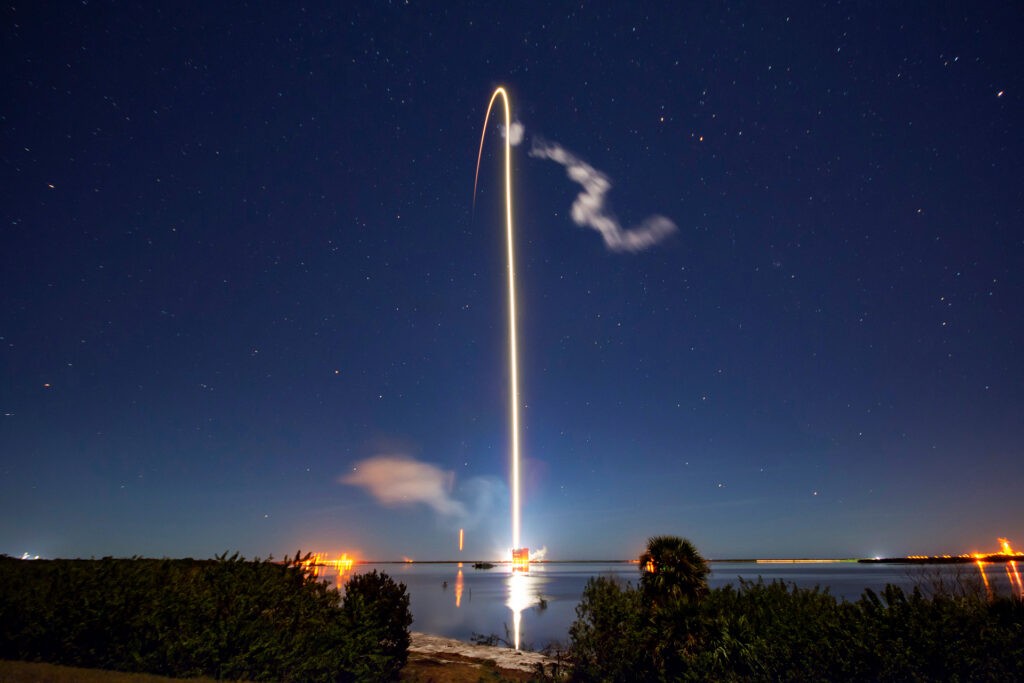 Rocket launching into the night sky above water, leaving a bright trail of light