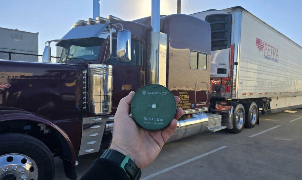 Hand holding a Surfact temperature monitoring device in front of a parked semi-truck